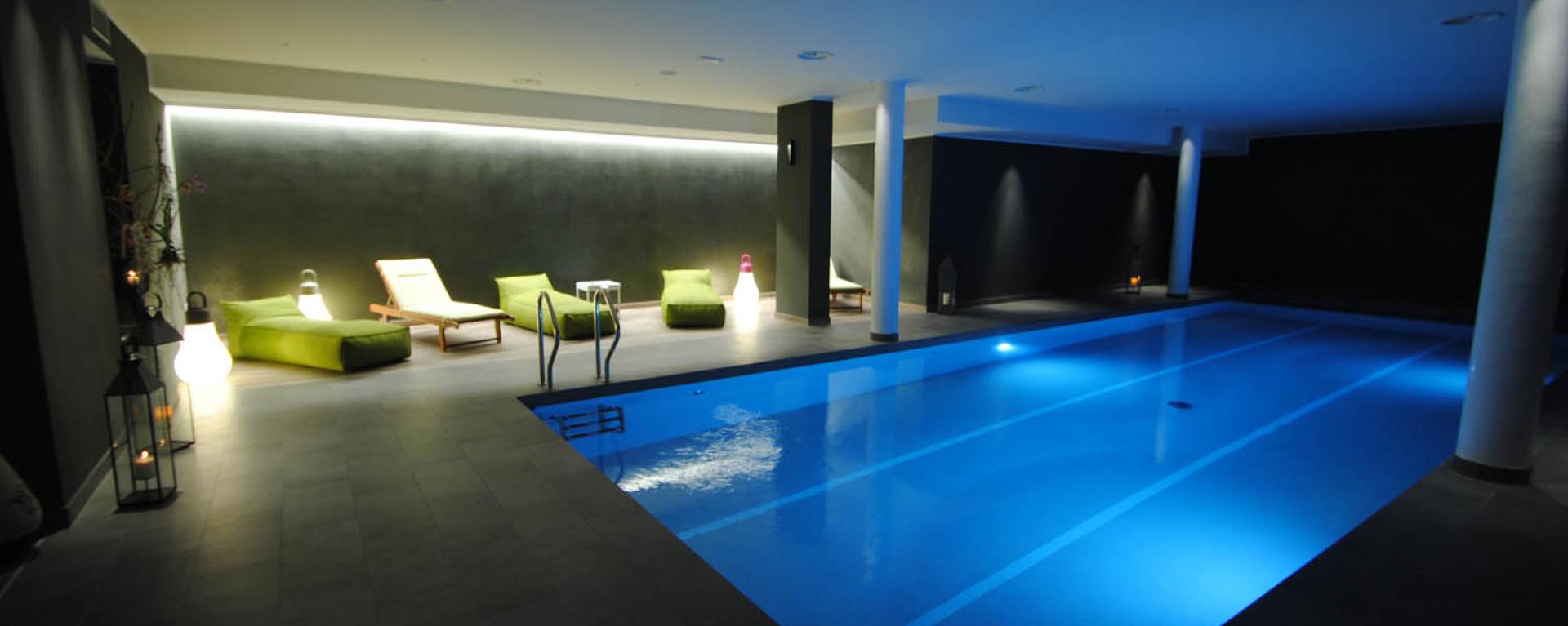 Piscina interna elegante con luci soffuse e comode sedute verdi per relax totale.