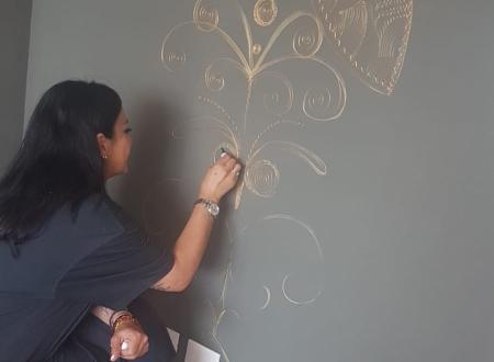 Donna disegna motivi dorati su parete grigia, creando un'atmosfera artistica e serena.